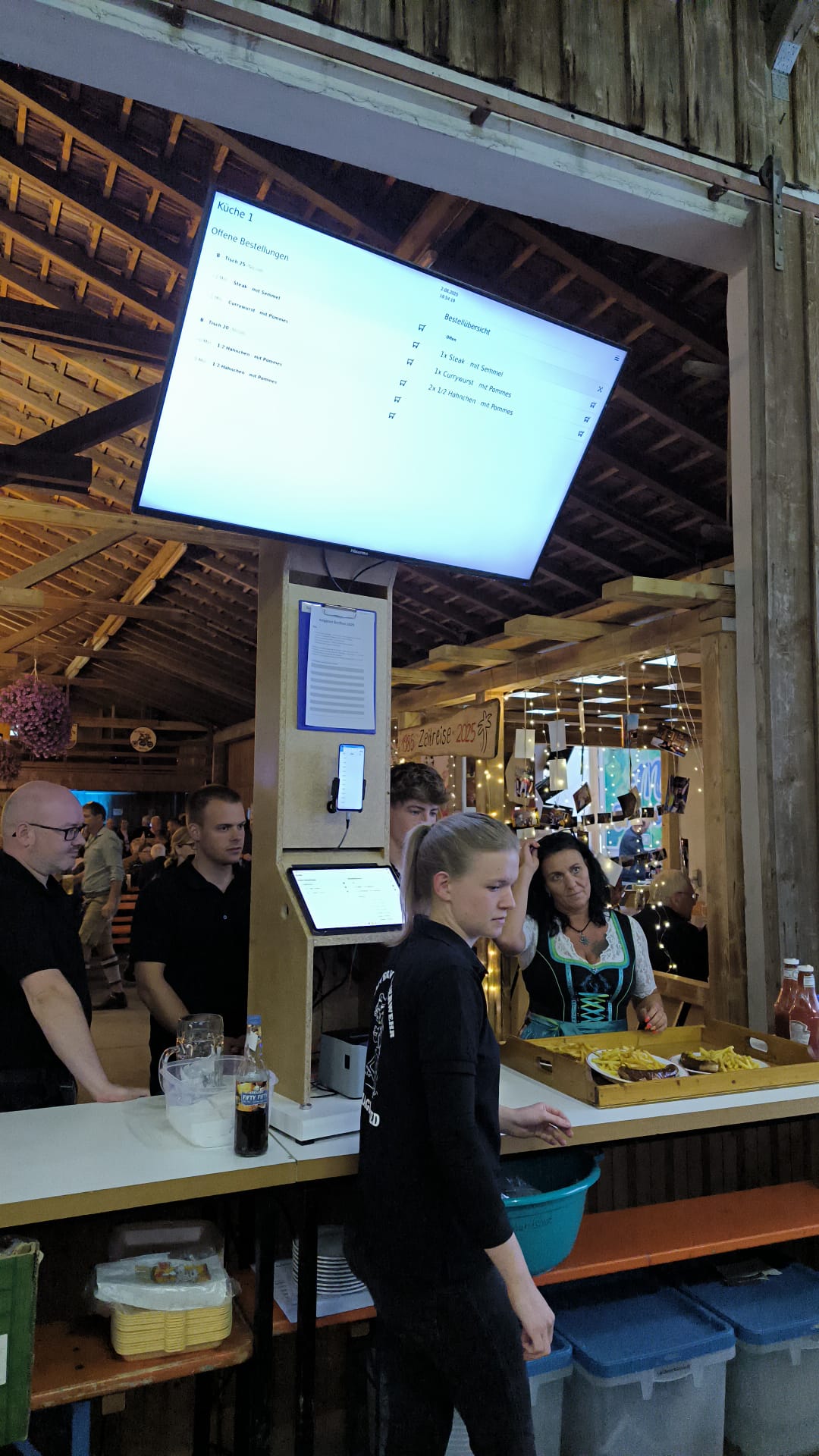 Großer Monitor des Bestellsystems Bassist über der Essensausgabe im Festzelt: offene Bestellungen pro Tisch und Bestellübersicht der Küche; darunter Tablet und Bondrucker, daneben fertige Teller. Großer Monitor des Bestellsystems Bassist über der Essensausgabe im Festzelt: offene Bestellungen pro Tisch und Bestellübersicht der Küche; darunter Tablet und Bondrucker, daneben fertige Teller.
