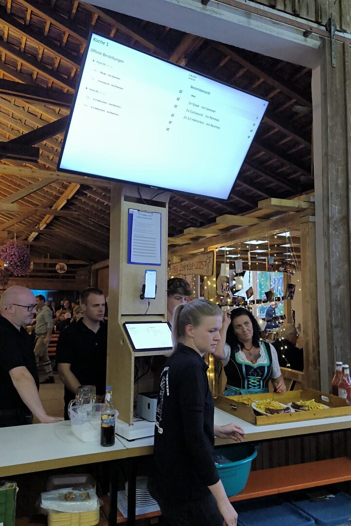 Essensausgabe bei einem Vereinsfest: Über der Ausgabe zeigt ein großer Monitor die offenen Bestellungen der Küche, darunter Tablet und Bondrucker; die Austräger holen die fertigen Teller mit dem Bon ab.